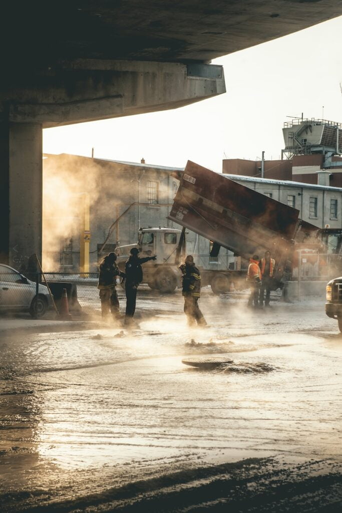 Blackcore Environmental - Workers and firefighters stand under a flyover near a tilted tipper lorry, with steam rising from the wet street in an industrial area.