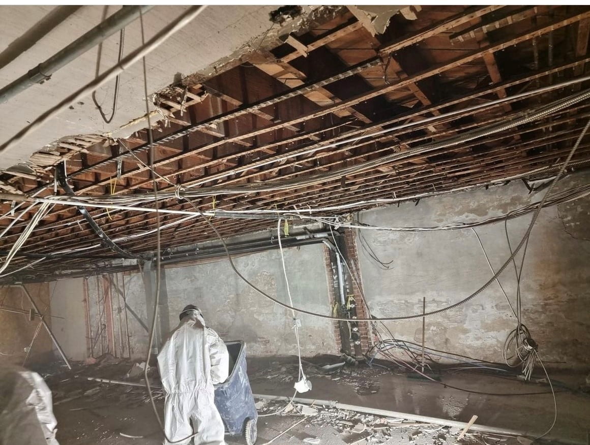 Blackcore Environmental - Two workers in protective suits clear debris beneath a partially demolished ceiling with exposed beams, wires, and damaged walls.