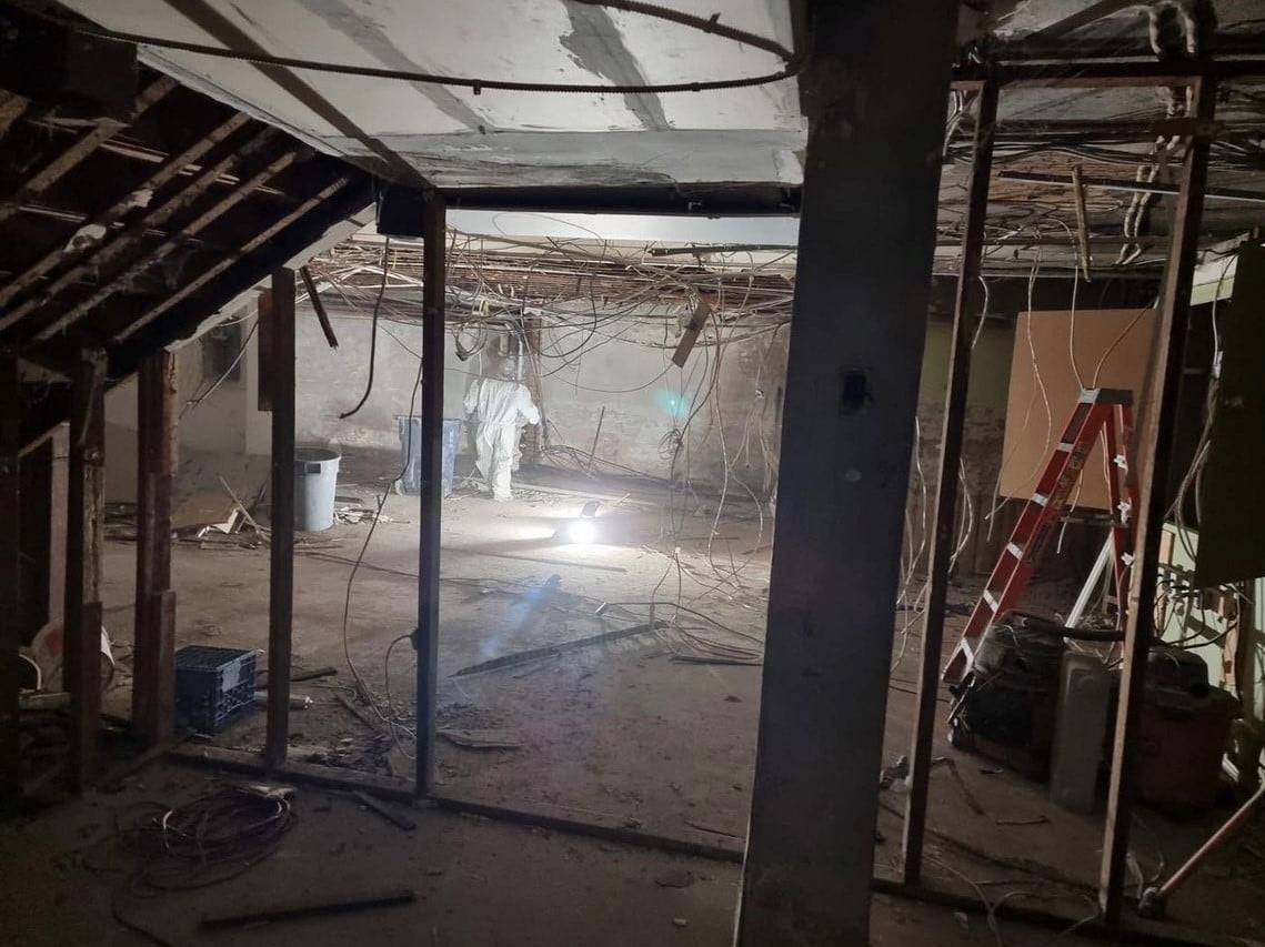 Blackcore Environmental - A dimly lit, partially demolished room with exposed wires and beams, construction debris on the floor, and a person in protective gear standing near a light.