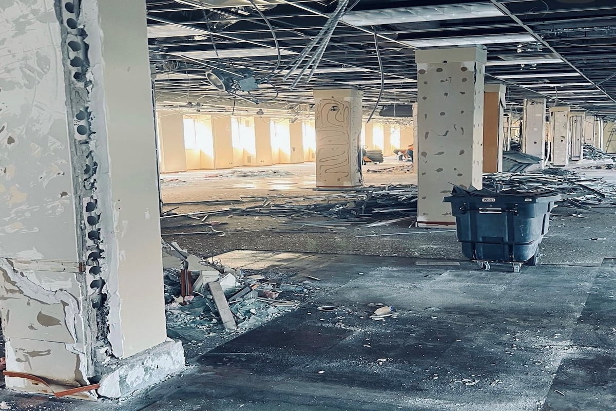 Blackcore Environmental - Large interior space undergoing demolition, with exposed pillars, debris scattered on the floor, and a black rubbish bin in the centre. Ceiling panels are missing, revealing wires and ducts.