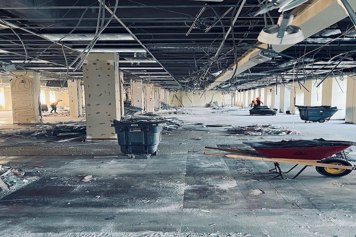 Blackcore Environmental - Large indoor space under demolition with exposed ceiling wires, debris on the floor, rubbish bins, and a red wheelbarrow in the foreground; workers are visible in the background.