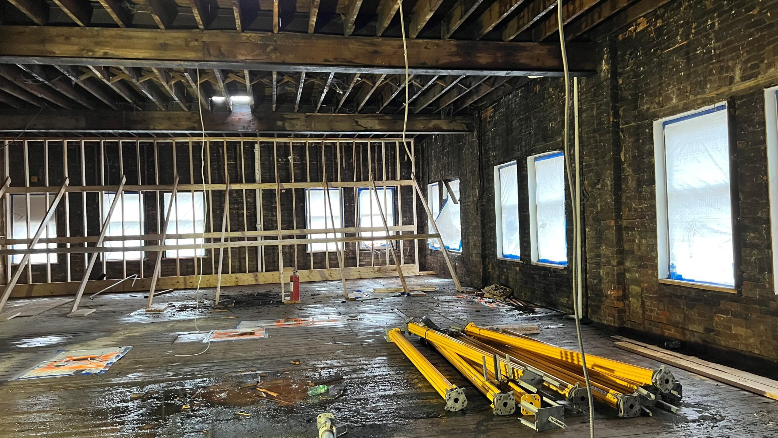 Blackcore Environmental - Interior of a large room undergoing refurbishment with exposed beams, unfinished walls, building materials, and windows covered with protective plastic.
