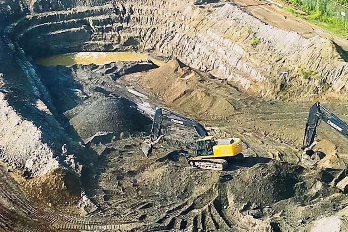 Blackcore Environmental - Two diggers dig and move earth in a large open-cast mine, with exposed soil layers and a shallow pool of water visible.