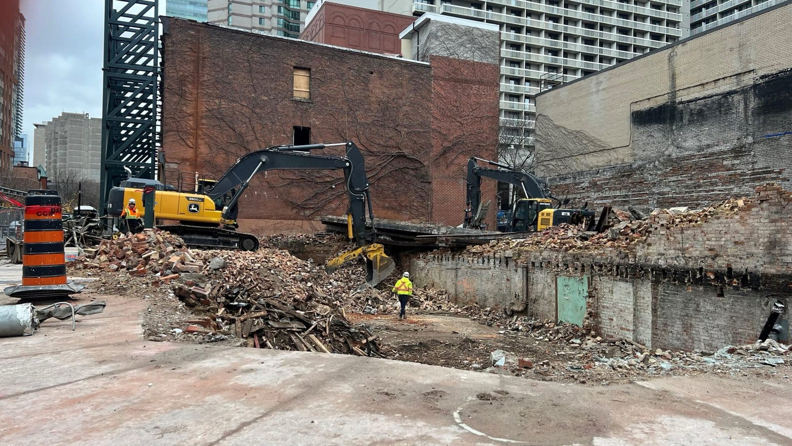 Blackcore Environmental - Two diggers clear rubble at a demolition site in a city, with a worker in a yellow jacket standing in the foreground among debris and brick walls.