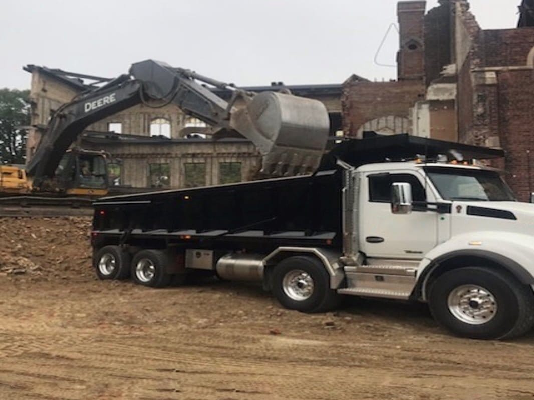 Blackcore Environmental - An excavator loads earth into the back of a white tipper lorry at a construction site with partially demolished brick buildings in the background.